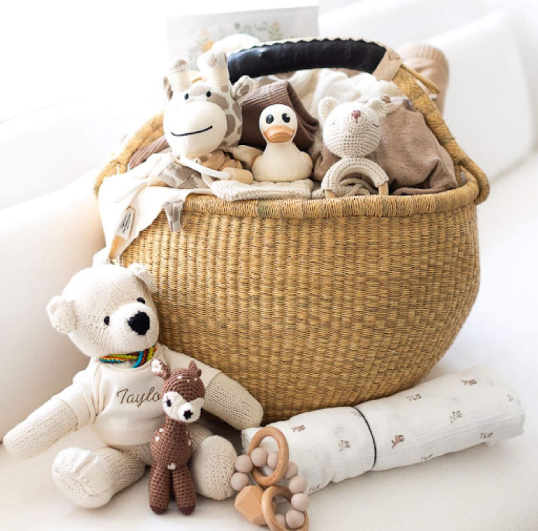 Woven straw baby gift basket filled with baby clothes and soft toys including a giraffe, duck, bears, and a fawn, with blanket and teether in front.
