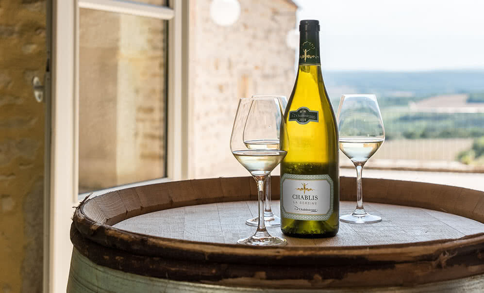 A bottle of 2020 La Chablisienne Chablis 1er Cru Mont de Milieu and three wine glasses on a barrel. 