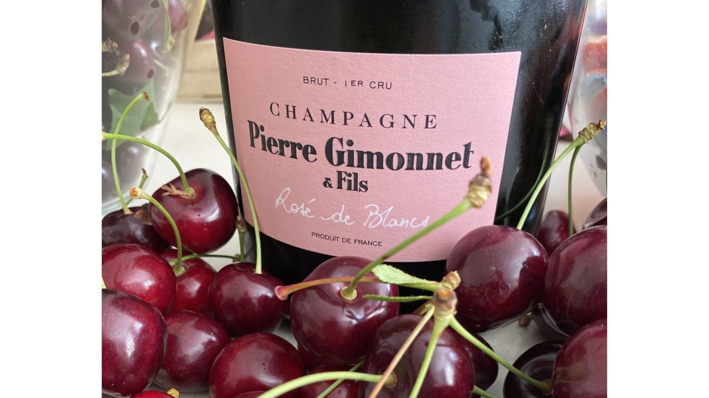 Champagne rosé bottle with pink label displayed among fresh cherries on a tabletop.