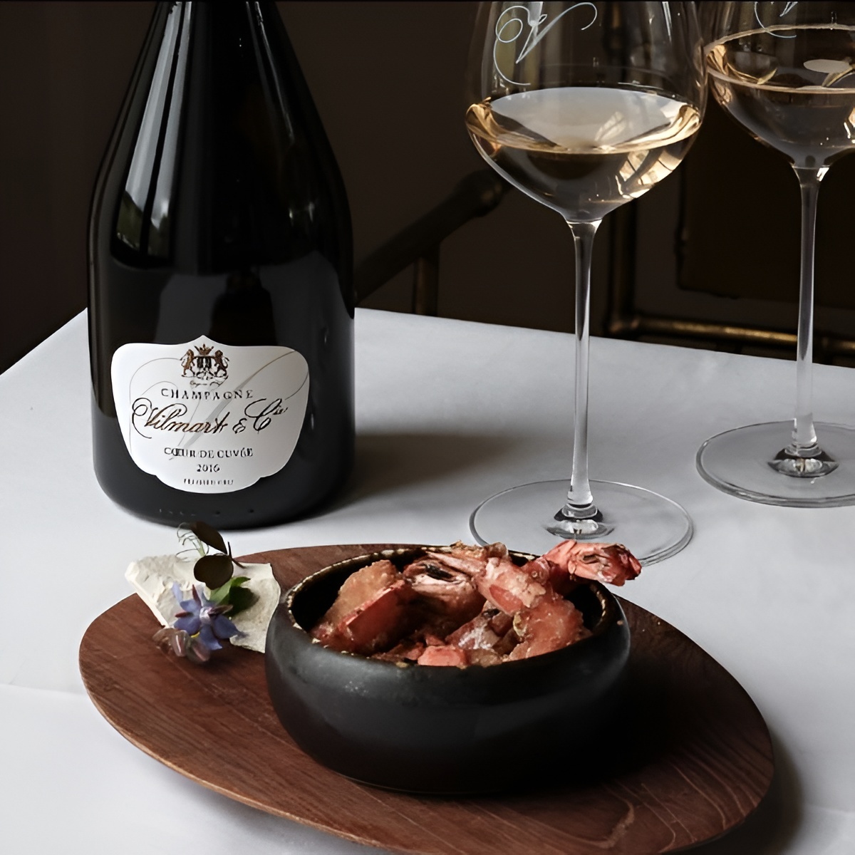A bottle of Champagne Vilmart & Cie next to two glasses and a small wooden plate holding a bowl of cooked shrimp.