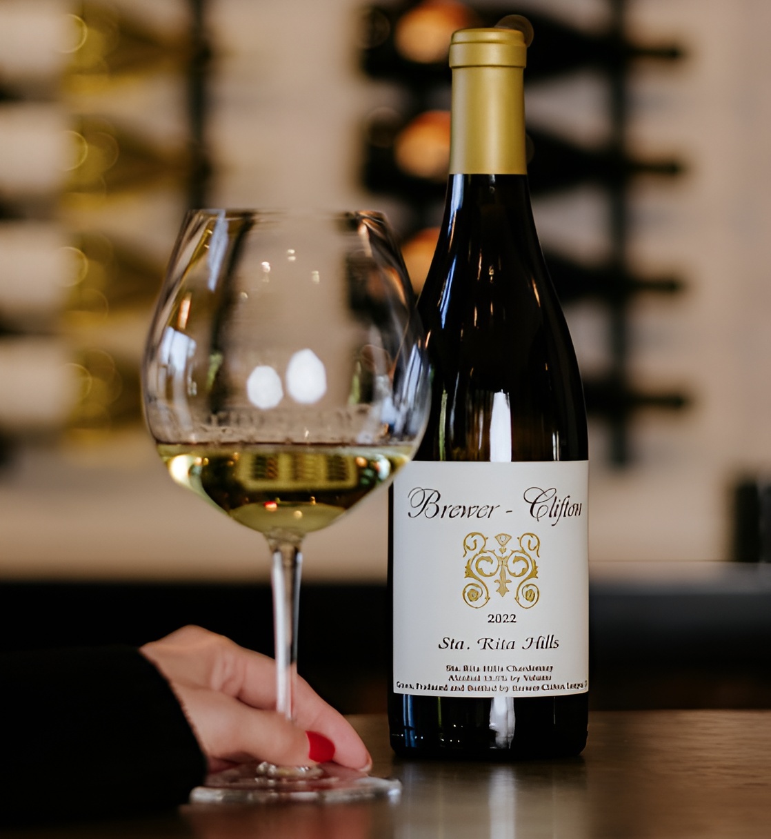 A hand holds a glass of white Chardonnay wine next to a bottle of Brewer-Clifton Sta. Rita Hills.