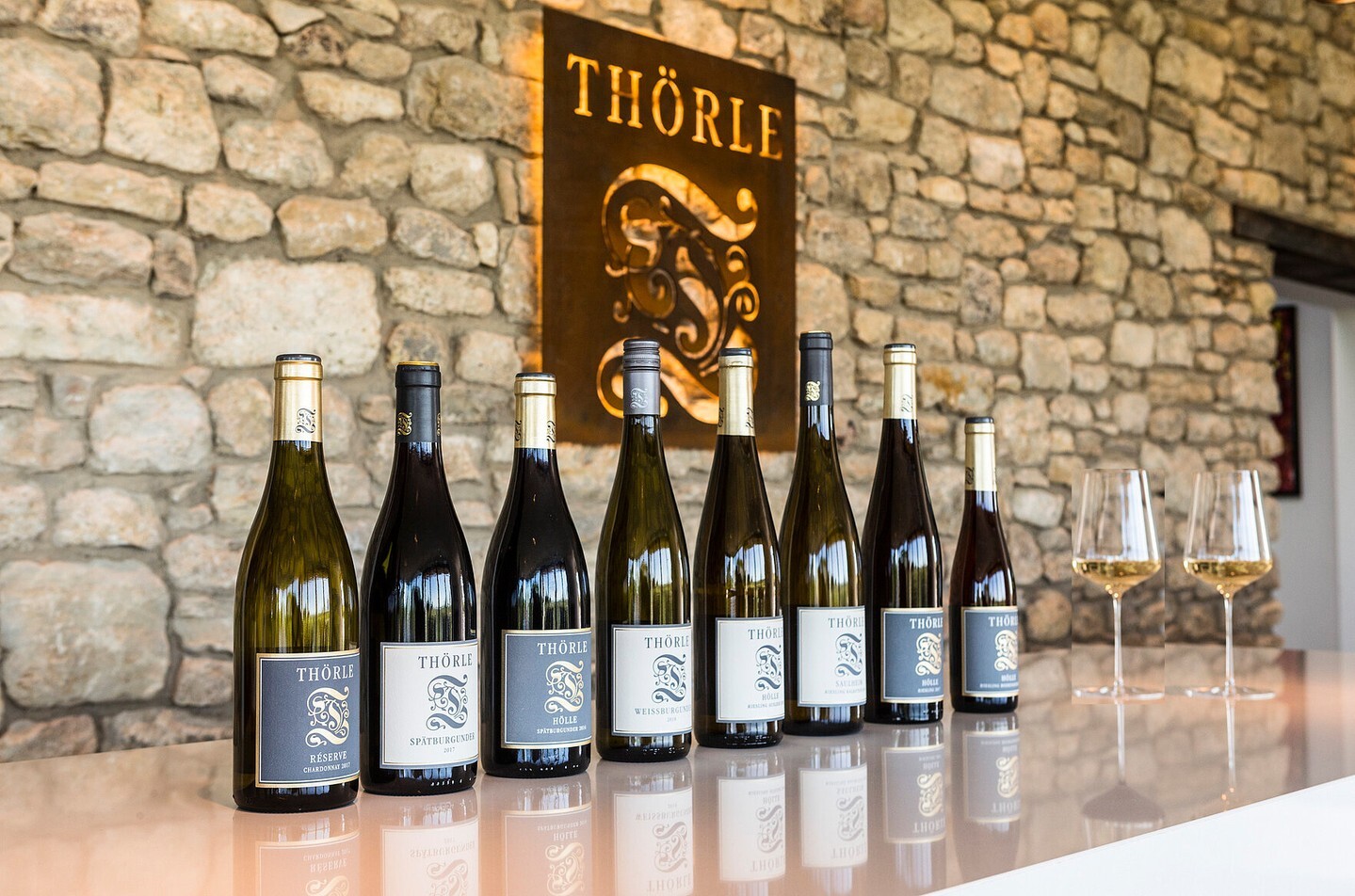 A row of eight assorted THÖRLE wine bottles and two empty wine glasses displayed against a stone wall with a branded sign.
