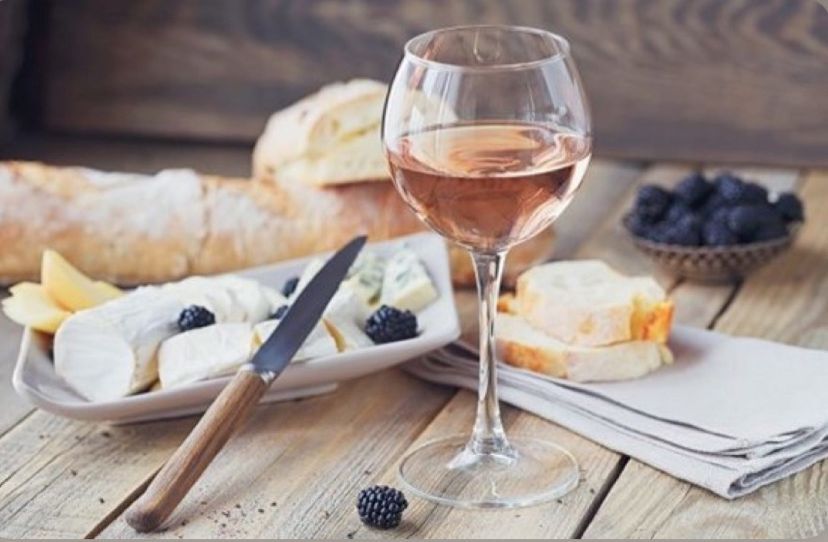 Glass of rosé wine with a cheese platter, fresh baguette, and blackberries on a rustic wooden table setting. 