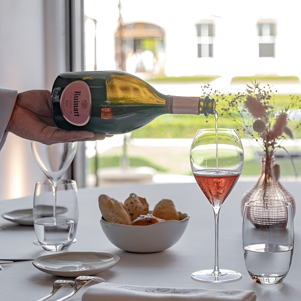 A bottle of Ruinart ros&eacute; is being poured into a glass at a bright, relaxed table setting.