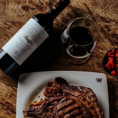 Grilled ribeye steak paired with a glass and a bottle of red wine on a rustic wooden table.