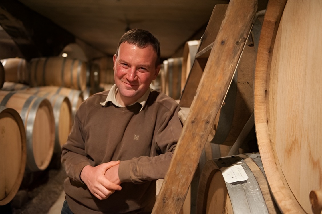  A smiling winemaker standing in a dimly lit cellar surrounded by rows of large wooden wine barrels.